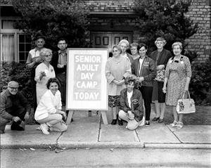 Twelve white people, men and women, mostly elderly stand and kneel in front of a brick building and around a sign